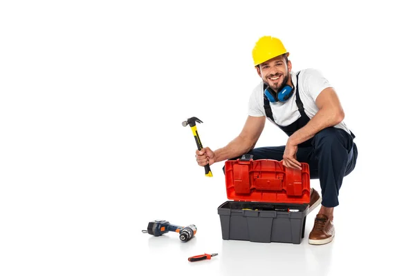 Operaio sorridente in uniforme che tiene martello vicino cassetta degli attrezzi e strumenti su sfondo bianco — Foto stock