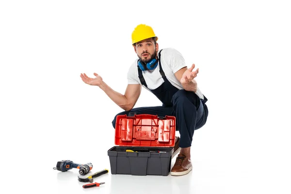Obrero confuso mostrando gesto de encogimiento cerca de herramientas y caja de herramientas sobre fondo blanco - foto de stock