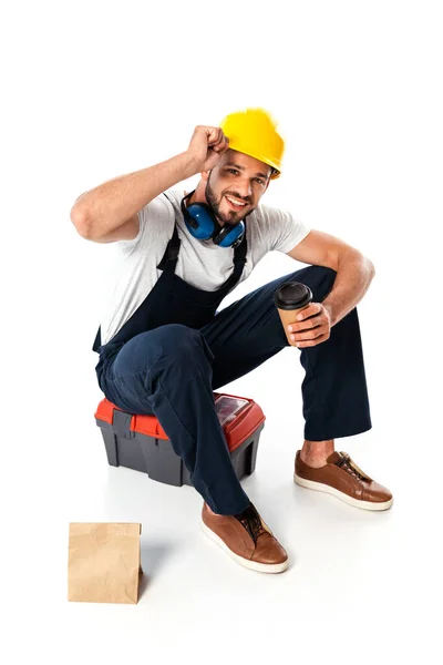 Рабочий-кузнец в форме и твердой шляпе держит кофе, чтобы пойти, сидя на инструментальной коробке рядом с бумажным пакетом на белом фоне — стоковое фото