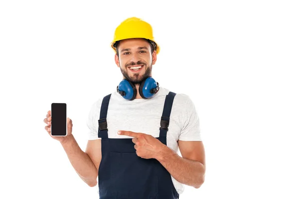 Trabajador sonriente en uniforme y casco protector apuntando con el dedo al teléfono inteligente aislado en blanco - foto de stock