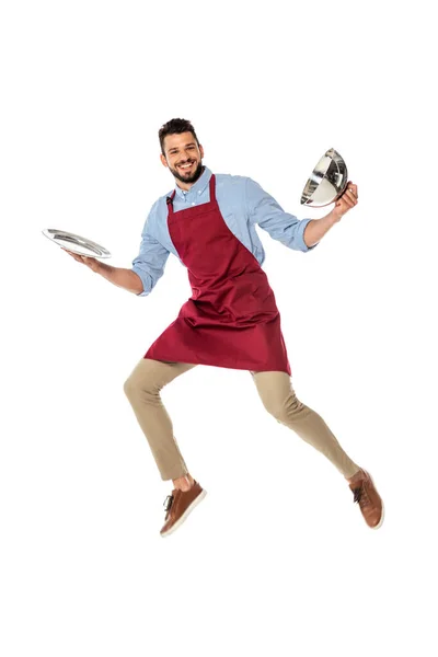 Camarero positivo en delantal sosteniendo bandeja y tapa del plato mientras salta aislado en blanco - foto de stock