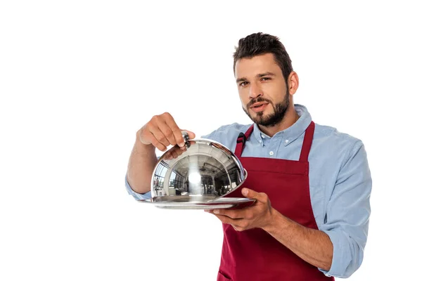 Handsome waiter in apron holding metal tray and dish cover while looking at camera isolated on white — Stock Photo