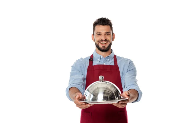 Усміхнений офіціант в фартусі, що показує металевий лоток і кришку посуду ізольовані на білому — стокове фото