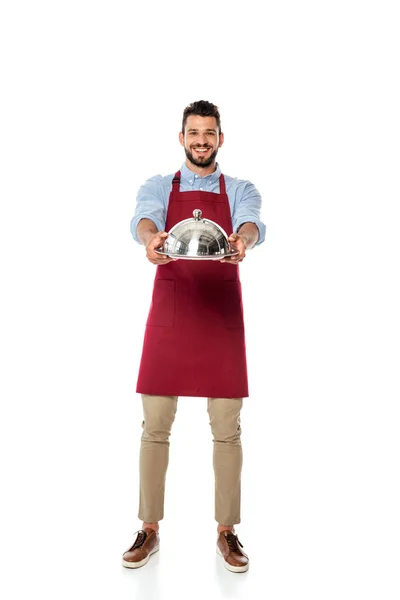 Camarero guapo sonriendo a la cámara y mostrando bandeja de metal y cubierta de plato sobre fondo blanco - foto de stock