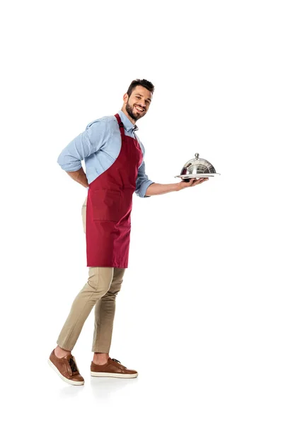 Бородатый официант в фартуке с металлической крышкой и подносом, улыбаясь в камеру на белом фоне — стоковое фото