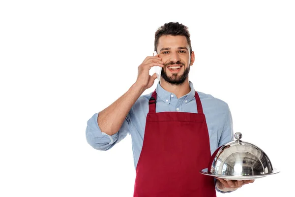 Smiling waiter talking on smartphone and holding tray with dish cover isolated on white — Stock Photo