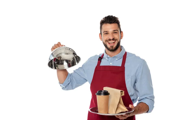 Cheerful waiter holding paper cup and bag on tray and dish cover isolated on white — Stock Photo