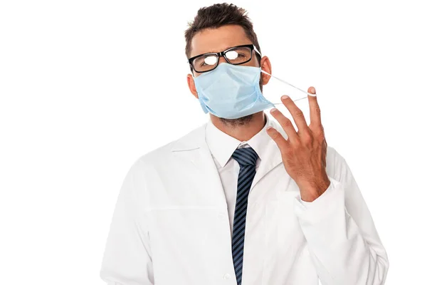 Young doctor in white coat holding strap of medical mask isolated on white — Stock Photo