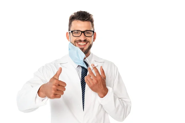 Sorridente medico mostrando pollice in alto e tenendo la maschera medica isolata su bianco — Foto stock