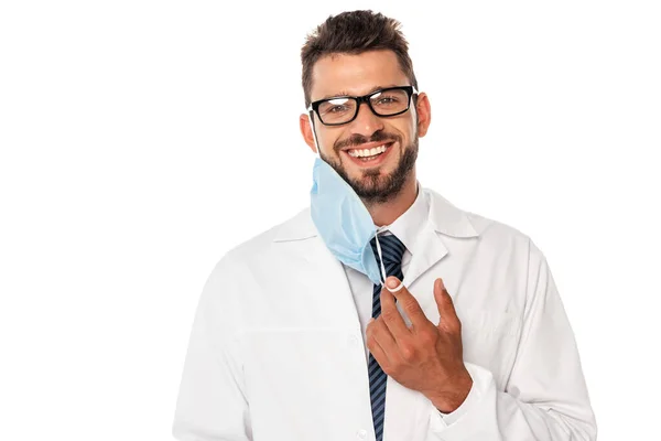 Beau médecin souriant à la caméra et tenant le masque médical isolé sur blanc — Photo de stock