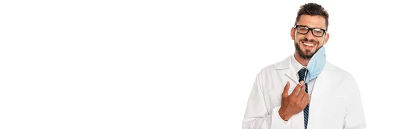Панорамний знімок красивого лікаря, який посміхається на камеру, тримаючи медичну маску ізольовано на білому — стокове фото