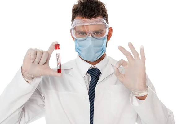 Médico en máscara médica sosteniendo tubo de ensayo con muestra de sangre y mostrando gesto aceptable aislado en blanco - foto de stock