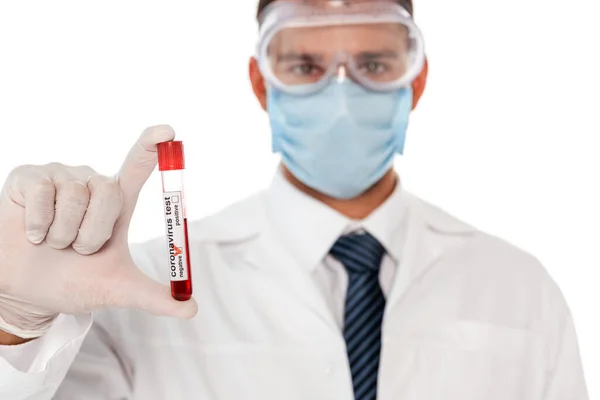 Focus selettivo del medico in maschera protettiva e occhiali tenuta provetta con campione di sangue e coronavirus lettering isolato su bianco — Foto stock