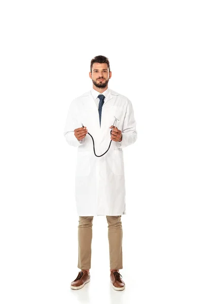 Молодой бородатый врач в белом халате держит стетоскоп изолирован на белом — стоковое фото