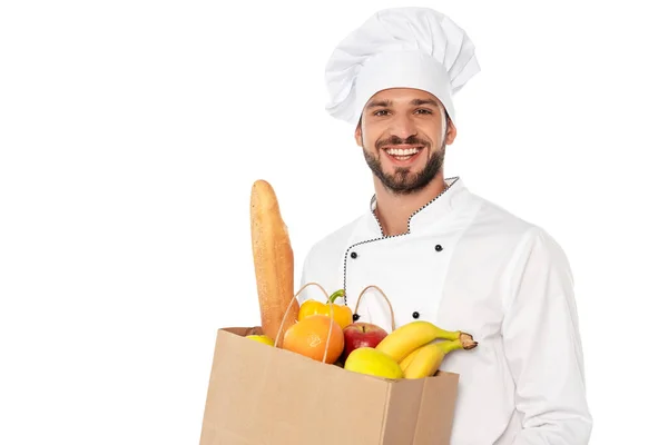 Handsome chef smiling at camera and holding shopping bag with baguette and fresh fruits isolated on white — Stock Photo