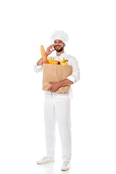 Chef souriant tenant un sac à provisions avec de la nourriture et parlant sur smartphone sur fond blanc — Photo de stock