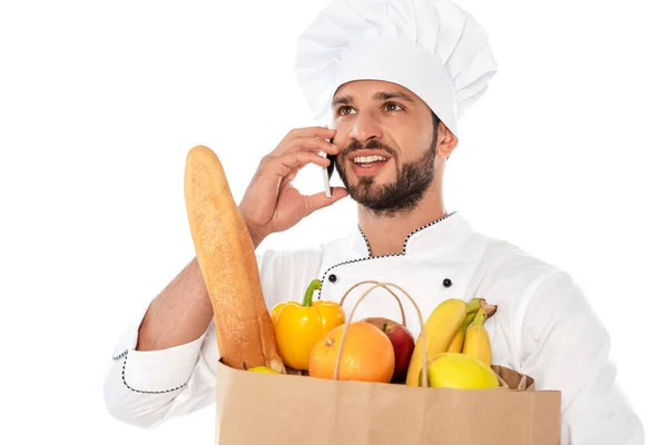 Cocinero sonriente hablando en el teléfono inteligente y sosteniendo bolsa de compras con alimentos aislados en blanco - foto de stock