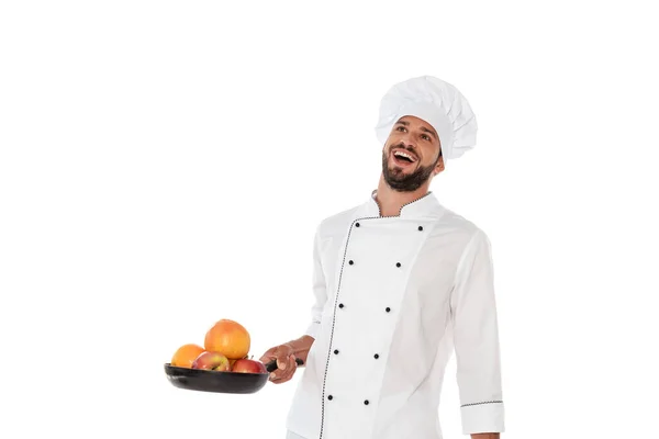Смеющийся шеф-повар в форме держит сковородку со свежими фруктами, изолированными на белом — стоковое фото