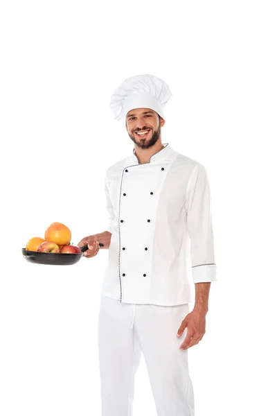 Handsome chef in uniform holding frying pan with ripe fruits and smiling at camera isolated on white — Stock Photo