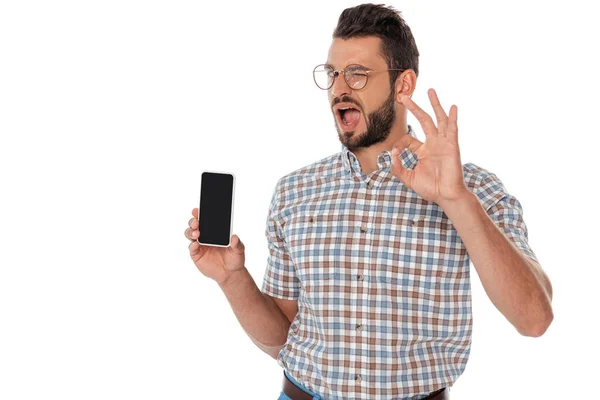 Nerd strizzando l'occhio e mostrando gesto ok tenendo smartphone isolato su bianco — Foto stock