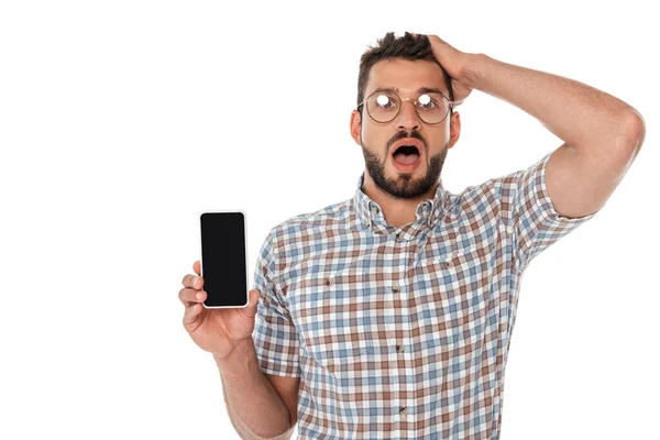 Nerd sorprendido con la mano cerca de la cabeza sosteniendo teléfono inteligente aislado en blanco - foto de stock