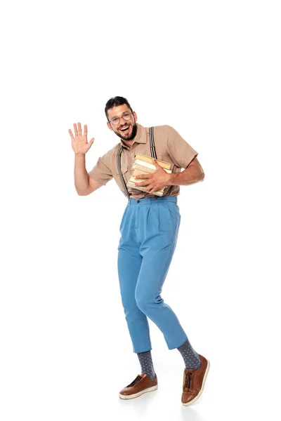 Cheerful nerd in suspenders holding books and waving hand at camera on white background — Stock Photo