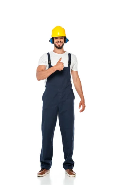 Guapo constructor en ropa de trabajo y hardhat sonriendo y mostrando el pulgar hacia arriba gesto sobre fondo blanco - foto de stock