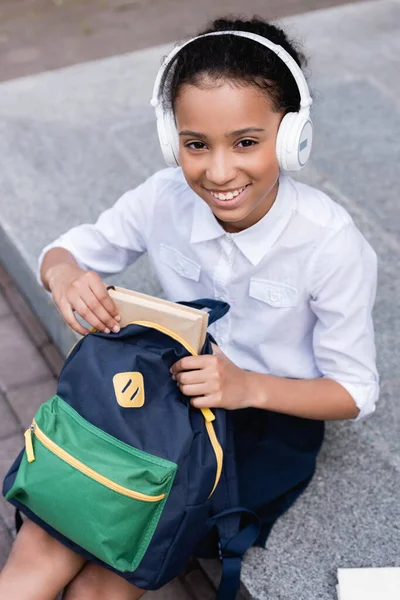 Colegiala afroamericana feliz en auriculares poner libro en la mochila - foto de stock