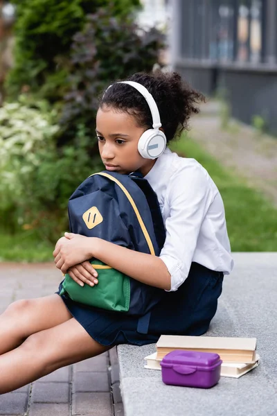 Sad african american schoolgirl with backpack and headphones sitting outside — Stock Photo