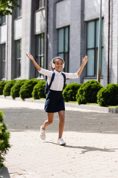 Happy african american schoolgirl with backpack and headphones walking on street with raised hands — Stock Photo