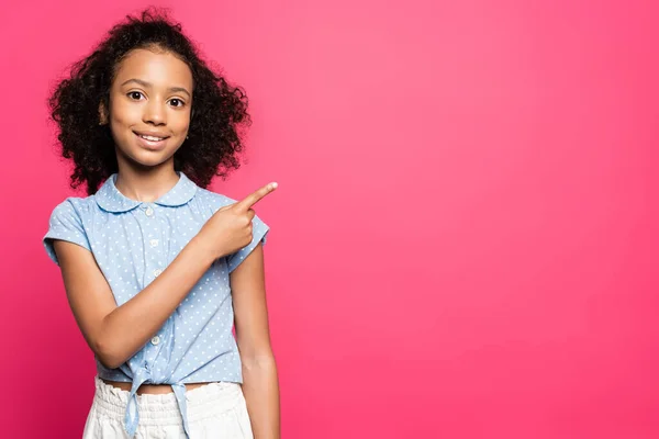 Sonriente lindo rizado africano americano niño señalando con el dedo a un lado aislado en rosa - foto de stock