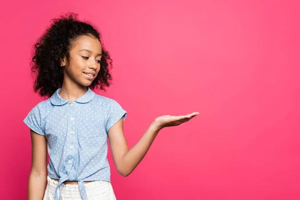 Lächelnd süße lockige afrikanisch-amerikanische Kind zeigt mit der Hand zur Seite isoliert auf rosa — Stockfoto