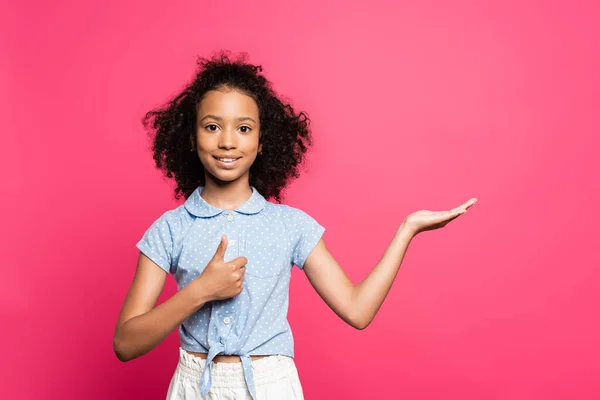 Lächelnd süße lockige afrikanisch-amerikanische Kind zeigt mit der Hand zur Seite und zeigt den Daumen nach oben isoliert auf rosa — Stockfoto