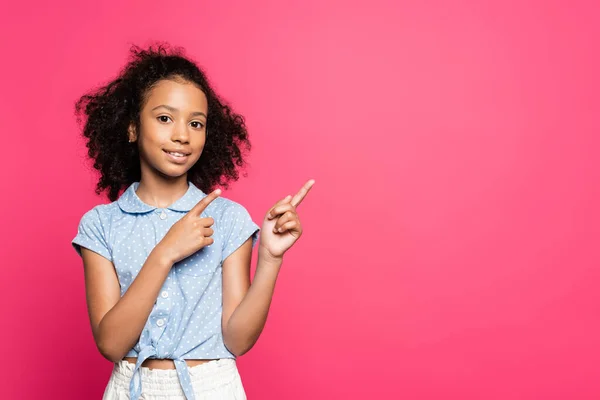Sonriente lindo rizado africano americano niño señalando con los dedos a un lado aislado en rosa - foto de stock