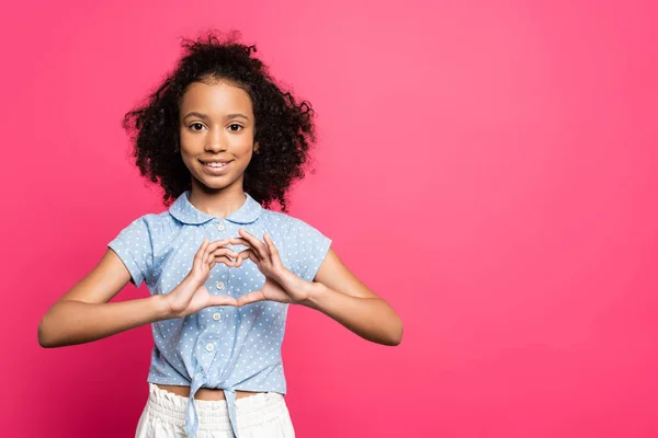 Lächelnd süße lockige afrikanisch-amerikanische Kind zeigt Herz mit Händen isoliert auf rosa — Stockfoto