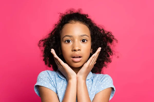 Chocado lindo rizado africano americano niño con las manos cerca de la cara aislado en rosa - foto de stock