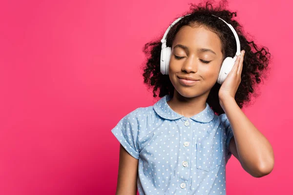 Smiling curly african american kid in headphones with closed eyes isolated on pink — Stock Photo