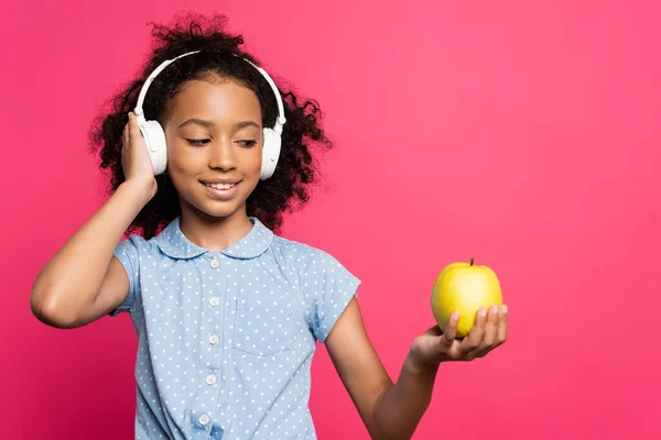 Sonriente rizado africano americano niño en auriculares celebración manzana aislado en rosa - foto de stock