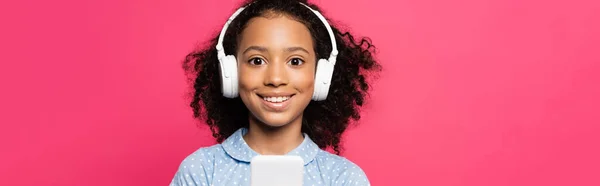 Lächelnd lockiges afrikanisch-amerikanisches Kind in Kopfhörern mit Smartphone isoliert auf rosa, panoramische Aufnahme — Stockfoto