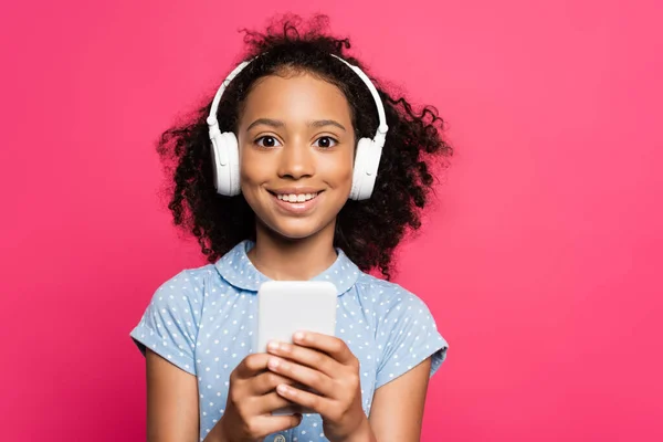 Sonriente rizado africano americano niño en auriculares usando teléfono inteligente aislado en rosa - foto de stock