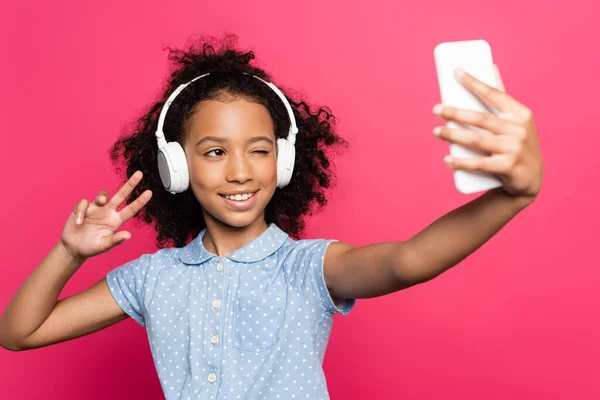 Foco seletivo de sorrir encaracolado criança americana africana em fones de ouvido mostrando sinal de vitória ao tomar selfie no smartphone isolado em rosa — Fotografia de Stock