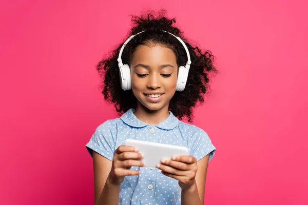 Sonriente rizado africano americano niño en auriculares usando teléfono inteligente aislado en rosa - foto de stock