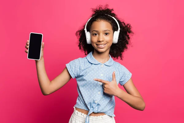 Smiling curly african american kid in headphones pointing at smartphone isolated on pink — Stock Photo