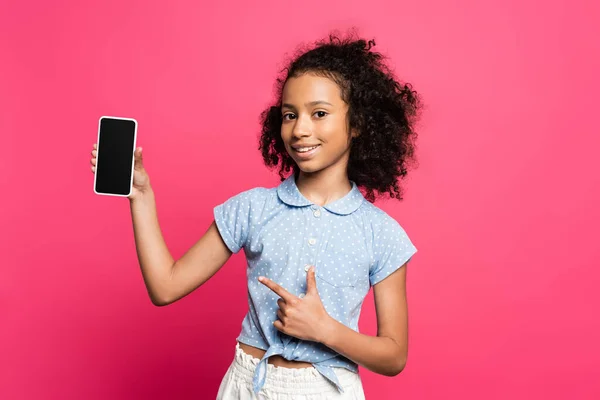 Smiling curly african american kid pointing at smartphone isolated on pink — Stock Photo