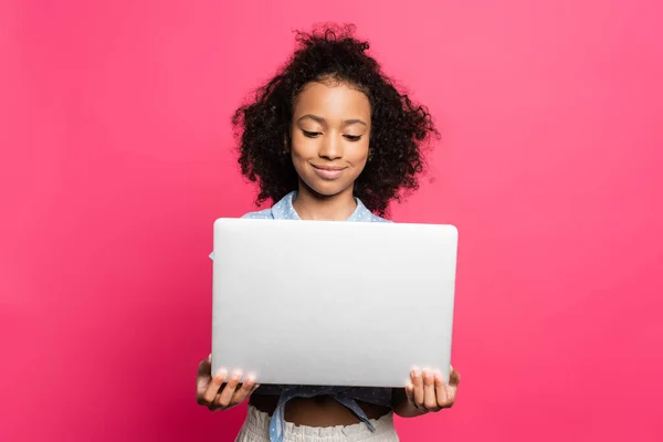 Lächelnd süße lockige afrikanische amerikanische Kind hält Laptop isoliert auf rosa — Stockfoto