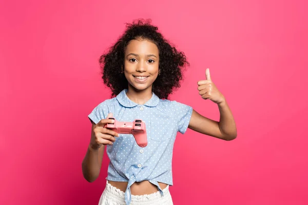 KYIV, UCRANIA - 9 de junio de 2020: sonriente niño rizado americano rizado con joystick que muestra el pulgar hacia arriba aislado en rosa - foto de stock