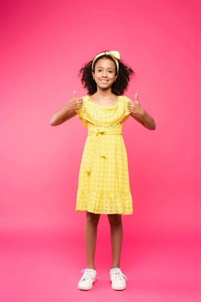 Full length view of smiling curly african american child in yellow outfit showing thumbs up on pink background — Stock Photo