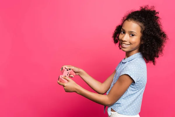 KYIV, UCRANIA - 9 de junio de 2020: sonriente lindo niño rizado afroamericano con joystick aislado en rosa - foto de stock