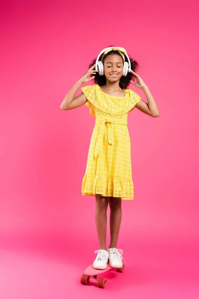 Full length view of smiling curly african american child in yellow outfit and headphones on penny board with closed eyes on pink background — Stock Photo
