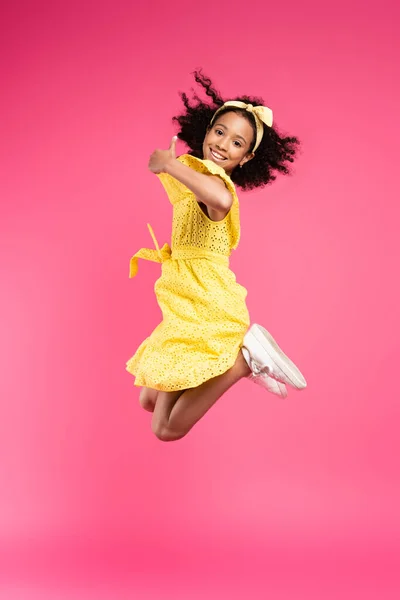 Vista laterale di felice riccio bambino africano americano in abito giallo salto e mostrando pollice su sfondo rosa — Foto stock